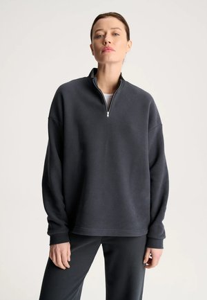 Kobieta z włosami zaczesanymi do tyłu, ubrana w ciemnoszarą bluzę z suwakiem do ćwiartki i pasujące spodnie na jednolitym jasnym tle.