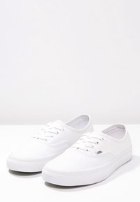 Para białych sneakersów low-top Vans z sznurówkami, górną częścią z płótna i gumowymi podeszwami na jednolitym białym tle.