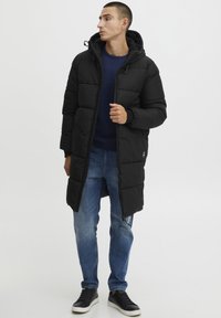 Sort puffet jakke med hætte, der har lynlåslukning, bred quiltet design og ribmede manchetter. Bæres over en navy sweater og jeans.