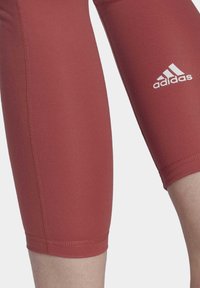 Adidas leggings pituutuneen polven pituuteen tummanpunaisesta kankaasta, joissa on litteät saumat ja valkoinen logo alaosassa, tarjoavat tiukan istuvuuden.