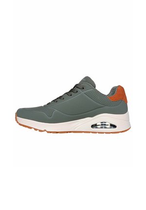 Zapatilla gris con parte superior lisa, acentos de ante marrón, ojales de metal y una unidad de amortiguación de aire visible. Suela de goma con banda de rodadura texturizada.