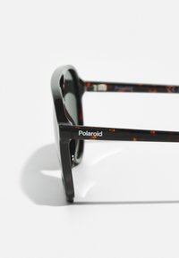 Ochelari de soare Polaroid cu un cadru glossy de culoare neagră și țestoasă, lentile închise la culoare și brand pe braț. Formă rectangulară.