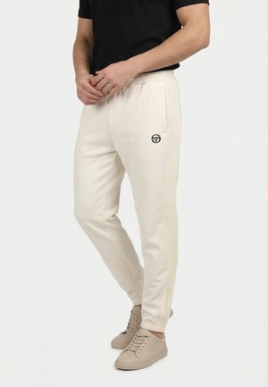 Hombre con pantalones jogger beige con rayas laterales y logo, usando zapatillas beige y camiseta negra, de pie frente a un fondo blanco liso.