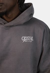 Szary hoodie z miękkiego materiału z przednią kieszenią i białym logo "CRYSTAL Paris" po lewej stronie klatki piersiowej.