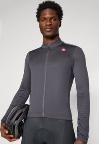 Maillot de cyclisme à manches longues en gris foncé, en tissu texturé avec une fermeture éclair sur le devant et un petit logo rouge sur la poitrine, tenu à côté d'un casque noir.
