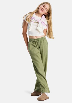 Młoda dziewczyna uśmiechająca się, ubrana w biały krótki t-shirt, zielone spodnie ze ściągaczem, beżowe buty wsuwane oraz różowo-paski sweter zawiązany na ramionach.