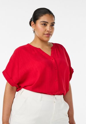 Femme aux cheveux foncés en chignon portant des boucles d'oreilles anneaux argentées, un chemisier rouge à boutons avec manches retroussées, et un pantalon blanc taille haute.