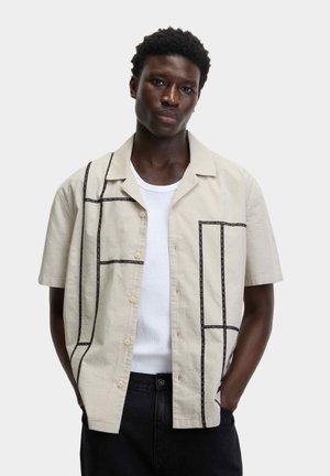 Korte mouwen beige shirt met zwarte geometrische stikpatronen, voorzien van een knopenlijst aan de voorkant en een ontspannen pasvorm, gelaagd over een witte tanktop.