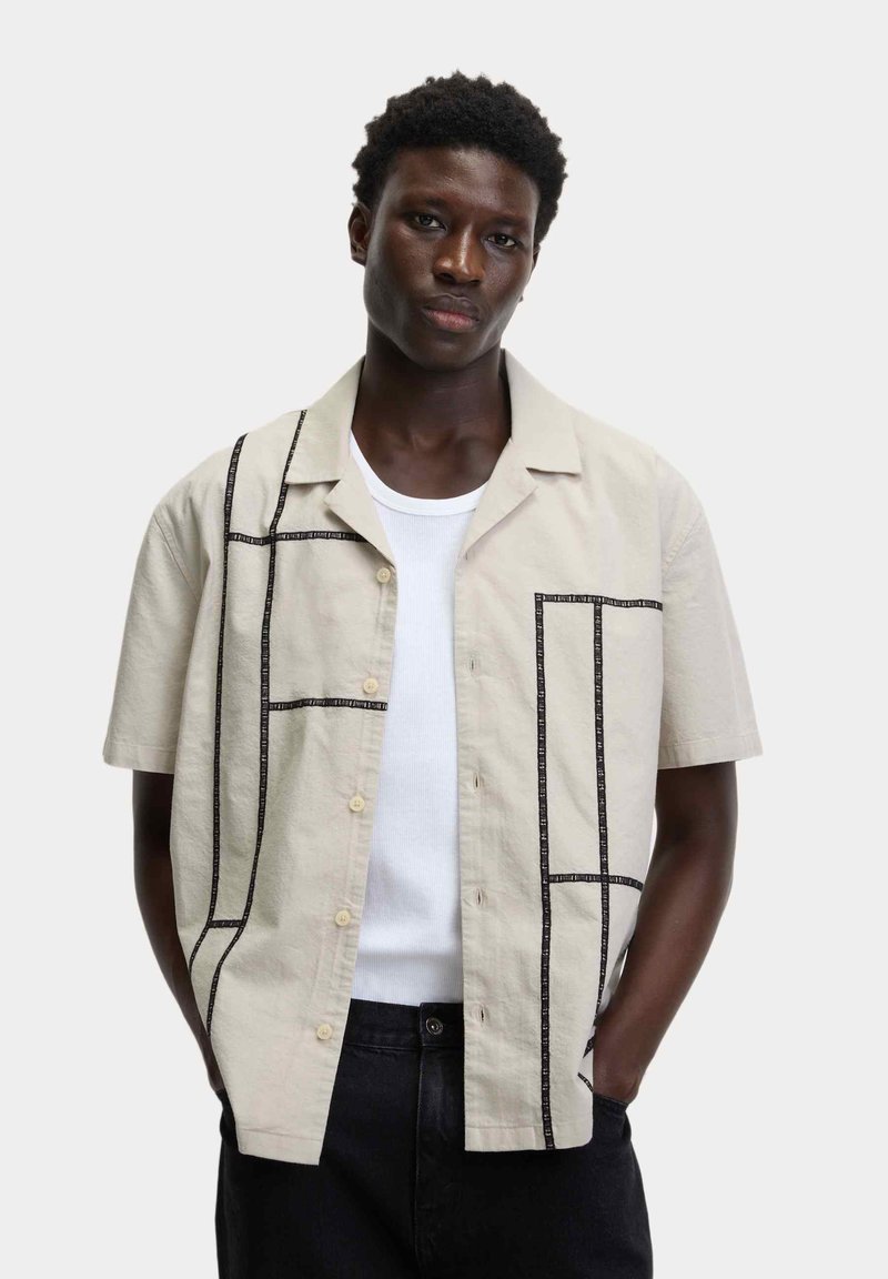 Kratší béžová košile s krátkým rukávem a černými geometrickými stehmi, s button-up přední částí a volným střih, překládající se přes bílý tílko.