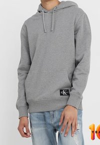 Šedá mikina s kapucí vyrobená z měkké tkaniny, s přední kapsou a nášivkou. Kombinováno s roztrhanými světle modrými džíny.