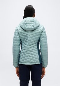 Person som har på seg en lyseblå quiltet jakke med hette, vist bakfra, mot en ensfarget hvit bakgrunn.