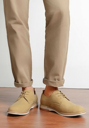 Chaussures décontractées à lacets de couleur marron clair portées avec un pantalon beige retroussé sur un sol en bois, sur fond blanc.