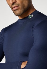 Top atlético de manga longa azul-marinho feito de um tecido liso e elástico. Apresenta um logo de jacaré verde na gola.