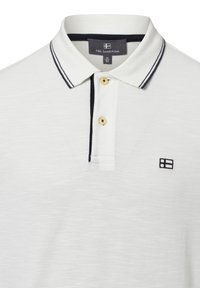 Weißes Poloshirt mit marineblauen Streifen am Kragen, ausgestattet mit zwei goldenen Knöpfen und einem kleinen schwarzen Logo auf der linken Brust. Strukturiertes Gewebe.