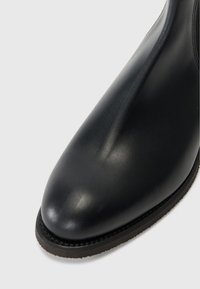Juoda odinė kulkšninių botą su apvaliu pirštu, lygiu tekstūru ir žemu, tamsiu guminio pado. Pabrėžta subtili siūlė aplink kraštą.
