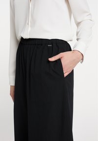 Zwarte, loszittende broek met een elastische tailleband en zijzakken. Het materiaal lijkt lichtgewicht, gecombineerd met een wit overhemd met knopen.