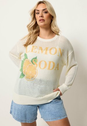 Femme blonde portant un pull en maille blanc avec texte jaune "LEMON SODA" et motif citron, associé à un short en jean bleu clair.