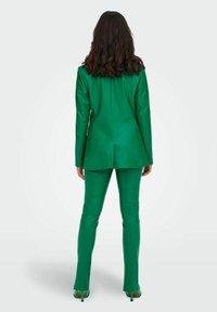 Completo in pelle verde con giacca strutturata e pantaloni aderenti. Le caratteristiche più rilevanti includono una texture liscia e orli dei pantaloni svasati.