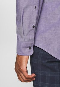 Fialová košile s dlouhým rukávem a jemnou texturou, s černými knoflíky a výšivkou růže u manžety. Kombinována s tmavě modrými kostkovanými kalhotami.