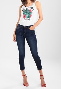 Witte tanktop met groen en roze bloemenprint, gecombineerd met donkerblauwe skinny jeans en rode sandalen met straps. Casual pasvorm, mouwloos ontwerp.