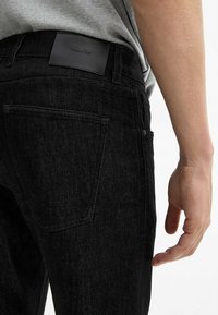 Jeans in denim nero con finitura testurizzata, con tasca posteriore e patch in pelle con il marchio sulla cintura, tenuti da una mano.