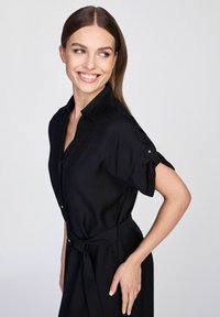 Czarna sukienka koszulowa z przeszyciem na guziki z przodu, krótkimi rękawami z podwiniętymi mankietami oraz pasującym paskiem w talii. Gładka faktura materiału.