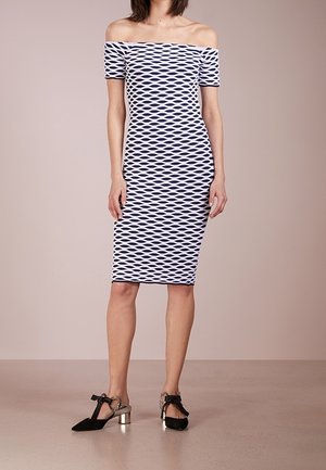 Femme portant une robe moulante longueur genou, épaules dénudées, avec un motif géométrique bleu et blanc et des escarpins noirs à bride à la cheville avec talons blocs métalliques.