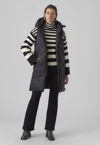 Gilet rembourré sans manches noir avec capuche, avec un design matelassé. Porté par-dessus un col roulé rayé noir et blanc et un pantalon noir.