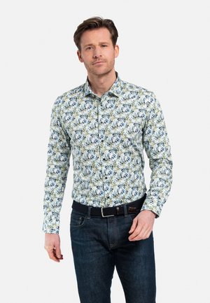 Chemise à boutons florale avec un motif de feuilles bleues et vertes sur un fond blanc, associée à un jean en denim foncé et une ceinture noire.