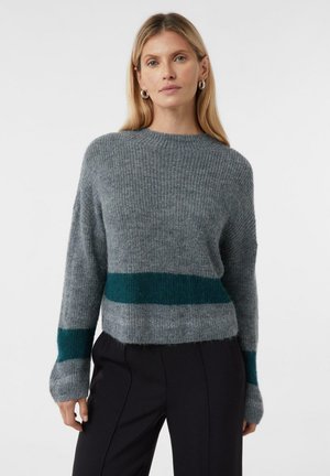 Woman wearing a gray knit sweater with dark green stripes on the lower sleeves and across the midsection, paired with black pants.