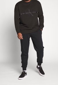 Czarna bluza z nadrukowanym logo, w zestawieniu z czarnymi spodniami dresowymi i czarnymi sneakersami. Prosty design oraz elastyczne mankiety.