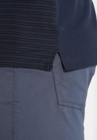 Gros plan sur l'ourlet d'une chemise à rayures bleu marine avec fente latérale, posé sur une poche de pantalon bleu foncé uni avec détails de couture.