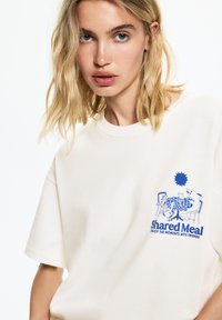 T-shirt en coton couleur crème à manches courtes, avec un imprimé graphique coloré d'animaux autour d'une table et le texte "Repas Partagé" à l'avant.
