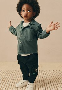 Veste en denim verte avec deux poches poitrine, associée à un jogging noir arborant un insigne et du texte. Des baskets blanches complètent la tenue.