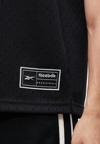 Μαύρη μπασκετική φανέλα Reebok με διατρητό ύφασμα και λογότυπο κοντά στο κάτω μέρος, δίπλα στο χέρι ενός ατόμου και μαύρα σορτς με λευκές ρίγες.