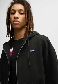 Felpa nera con zip, caratterizzata da un patch blu con il logo "HUGO" sul lato sinistro del petto. Indossata sopra una t-shirt nera con testo rosa.