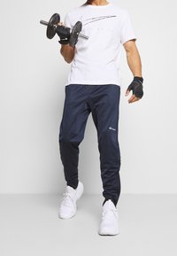 Vit T-shirt med kort ärm från Nike med svart grafik, marinblå träningbyxor, svarta vikterhandskar och vita sneakers.