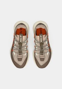 Chaussures de sport beige et marron avec une tige texturée, des accents orange et un système de laçage unique. Elles disposent d'une semelle en caoutchouc robuste.