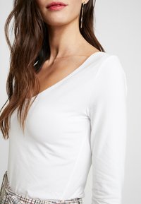 Weißes Langarmoberteil mit V-Ausschnitt. Weicher Stoff, figurbetonte Silhouette und sichtbare Nähte. Das Model trägt einen karierten Rock und auffällige Ohrringe.
