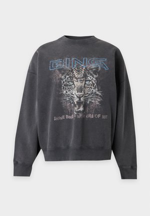 Femme portant un sweat-shirt oversize foncé avec un motif léopard et un texte bleu, associé à un jean droit bleu et des chaussures noires pointues.