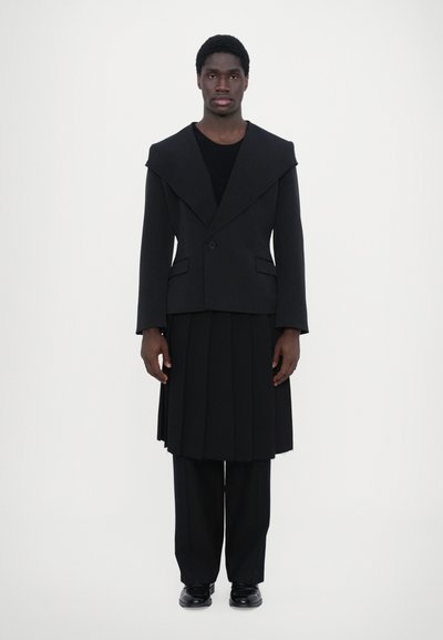 Schwarzer strukturierter Blazer mit breitem Kragen, kombiniert mit einem schlichten schwarzen Rock und schwarzen Hosen. Glatter Stoff, taillierter Schnitt, minimalistische Akzente.