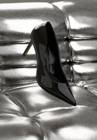 Scarpa con tacco alto in pelle verniciata nera, dalla punta affusolata e tacco sottile, posizionata su uno sfondo metallizzato argento lucido.