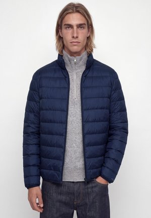 Jeune homme aux cheveux blonds mi-longs porte une veste matelassée bleu marine sur un pull gris zippé et un jean foncé, debout devant un fond blanc.