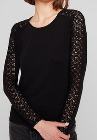 Schwarzes Langarmshirt mit aufwendigen Spitzenverzierungen an den Ärmeln, glatter Textur, Rundhalsausschnitt und taillierter Passform.