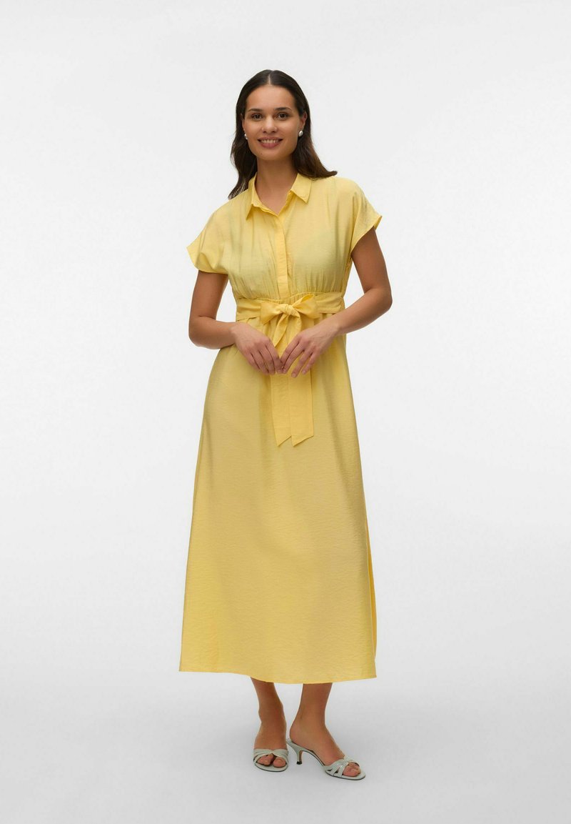 Kvinde står og smiler iført en gul midikjole med korte ærmer, et knyttet taljebælte og lyse sandaler med hæl.