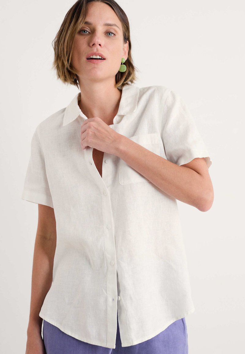 Camicia bianca di lino a maniche corte con bottoni frontali, colletto e una tasca sul petto, abbinata a pantaloni lavanda. Tessuto liscio.
