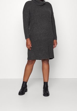 Osoba ubrana w długą, ciemnoszarą dzianinową sukienkę do kolan z długim rękawem oraz czarne botki, stojąca na jednolitym jasnym tle.