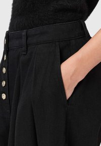 Pantalones plisados negros con cintura alta, con acentos de botones dorados en la parte frontal. El material parece texturizado y tiene bolsillos laterales.