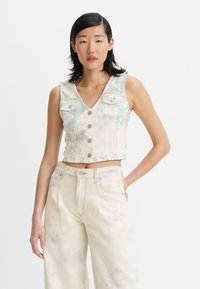 Cropped denim vest in light beige with blue tie-dye pattern. Features a V-neck, button closure, and two chest pockets. Paired with beige wide-leg jeans.