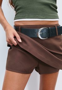 Bruine shorts van gladde stof, gecombineerd met een brede zwarte riem met een ronde zilveren gesp. Boven de shorts wordt een groene ribbeltop gedragen.